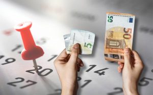 Mani che tengono una banconota da 5 e una da 50€. Calendario sullo sfondo